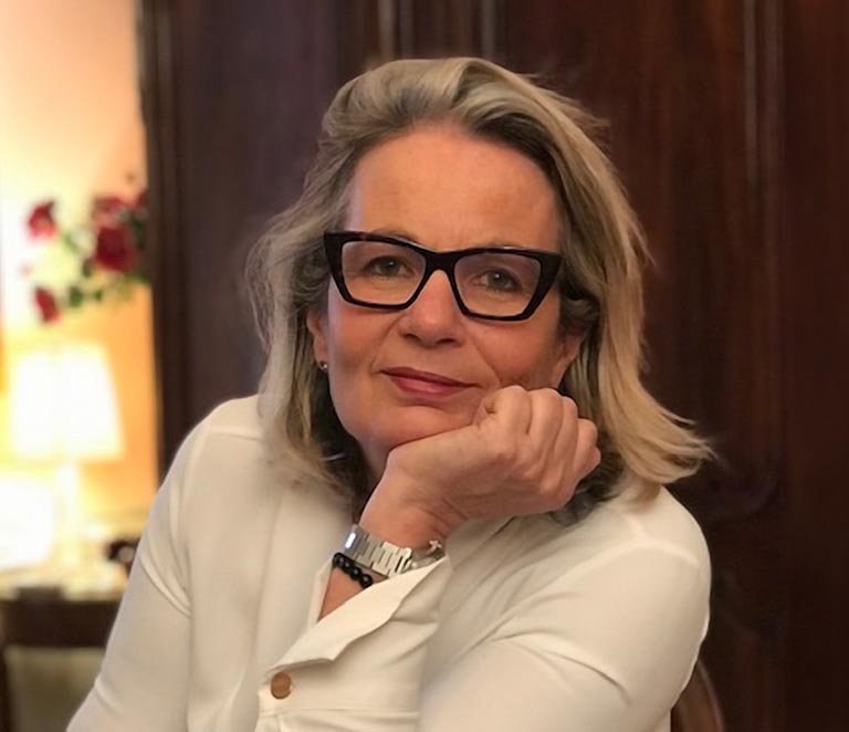 Delphine Faure seated indoors, wearing dark-framed glasses and a white top, resting a hand beneath the chin.